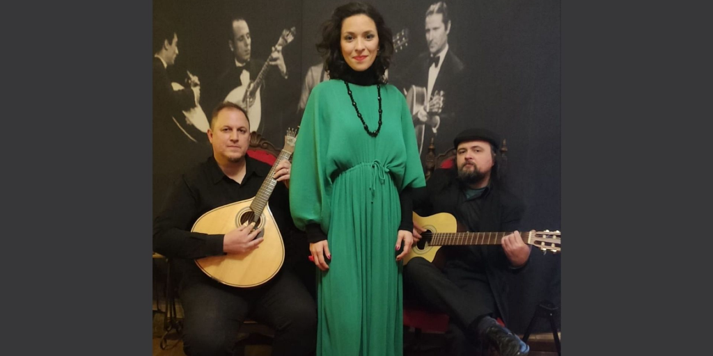 Imagem do grupo, dois homens brancos com instrumentos musicais e a cantora ao centro, branca de cabelos pretos curtos