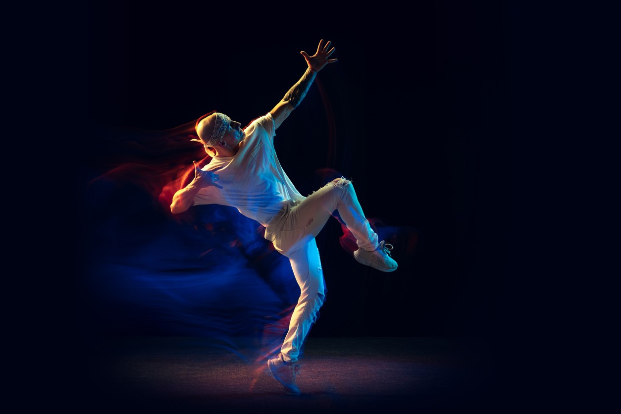 imagem de dançarino, branco careca, usando bandana e roupas brancas, em movimento se equilibra na perna esquerda enquanto ergo joelho direito e braços para o céu.