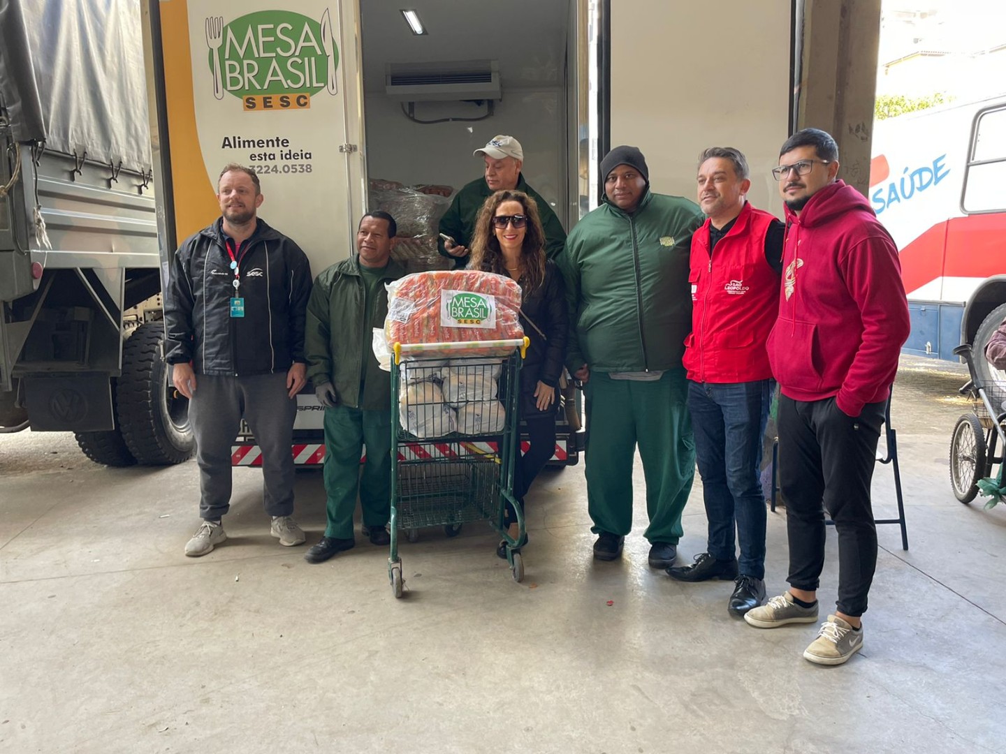 sete pessoas das equipes Unidade Sesc São Leo e Mesa Brasil posam em frente ao caminhão com um carrinho de mercado contendo sacas de alimentos