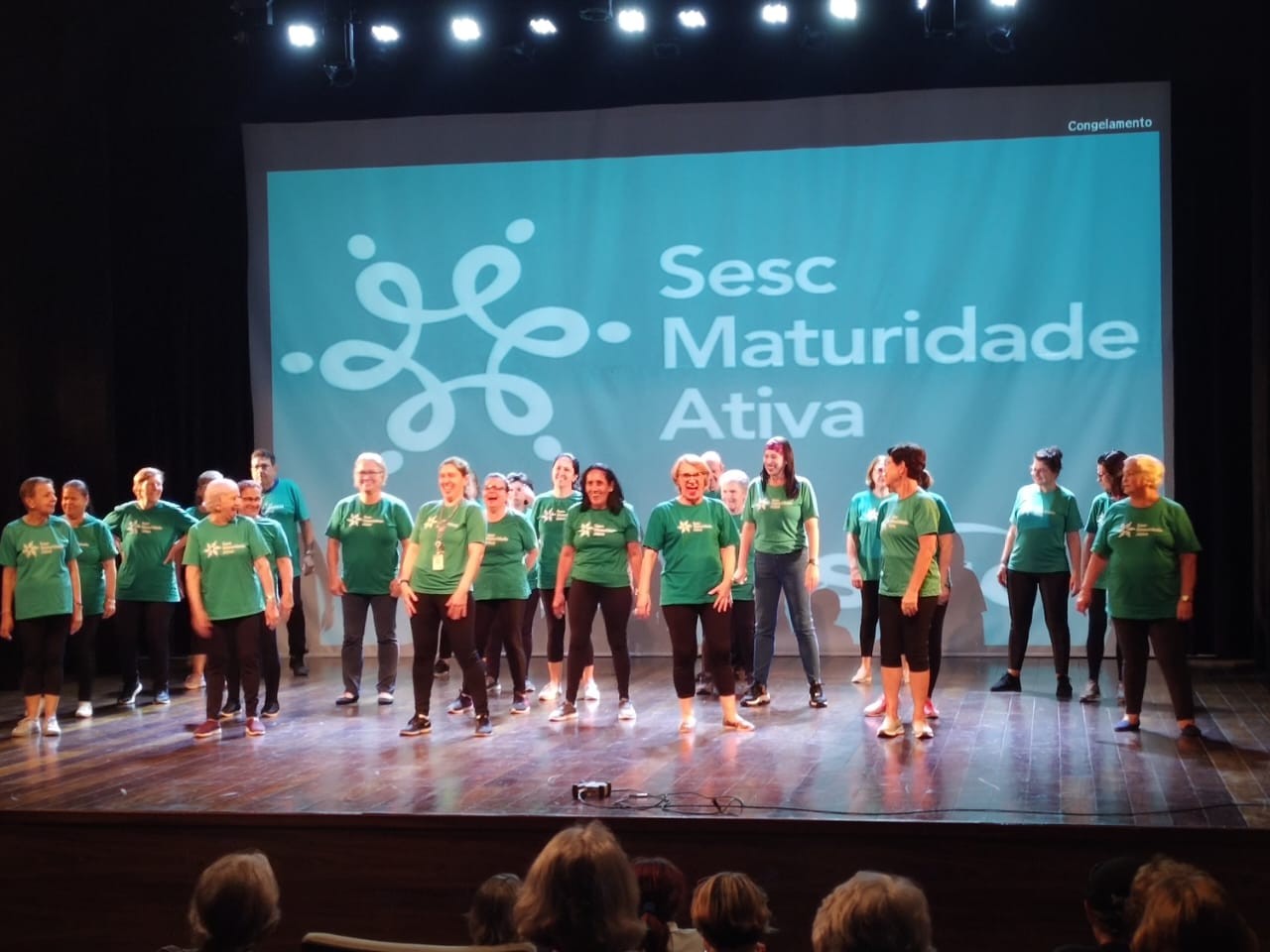 Imagem de 22 participantes da maturidade ativa de canoas sobre o palco do teatro com fundo de palco apresentando a logo e o nome "Sesc Maturidade Ativa"