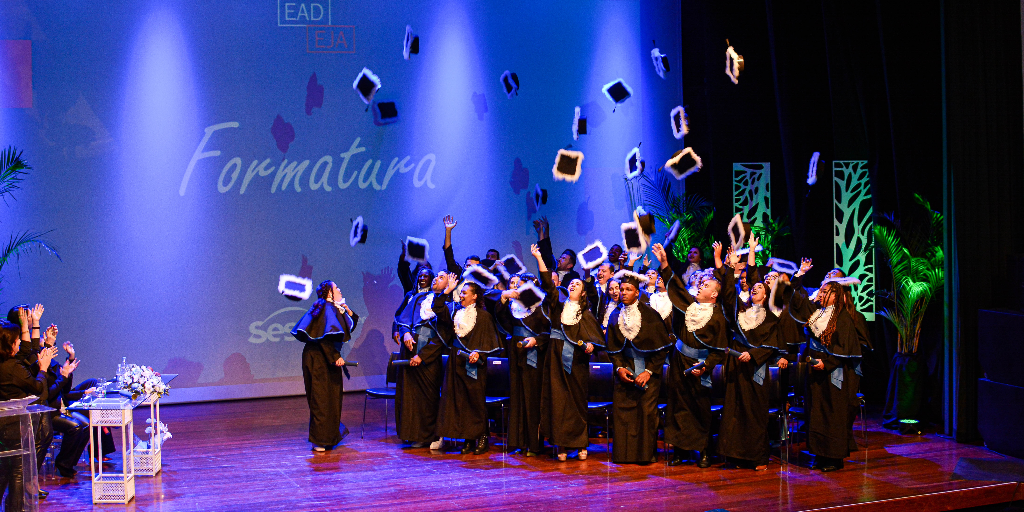 Grupo de estudantes de toga em cima do palco, lançando os capelos para o ar