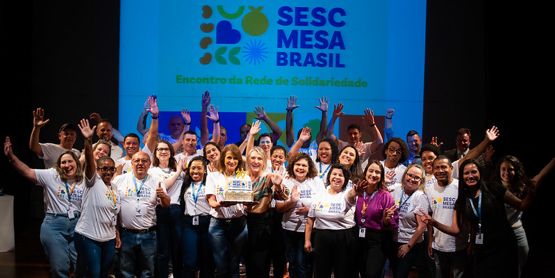 Grupo de pessoas vestindo a camiseta branca do Programa Mesa Brasil com a logo do programa projetada ao fundo