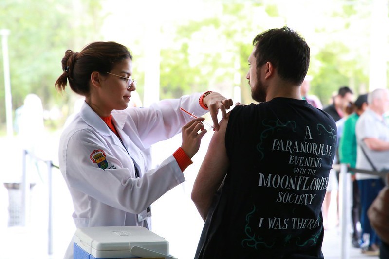 Homem de camiseta preta sendo vacinado por médica em uniforme branco