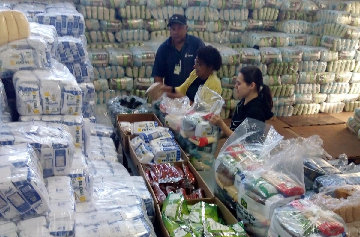 Três pessoas montando cestas básicas em armazém repleto de alimentos