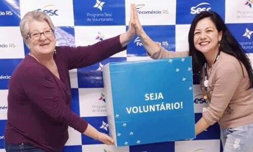 Duas mulheres tocando as mãos uma da outra em cima e segurando entre elas uma caixa azul com letras brancas dizendo "seja voluntário"