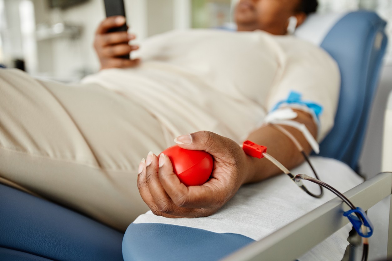 Pessoa de branco doando sangue enquanto aperta uma bola vermelha e mexe no celular