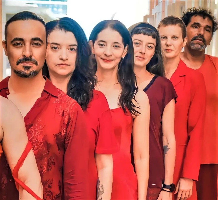 Foto mostra integrantes da performance teatral "Nós", todos alinhados e vestidos em vermelho.