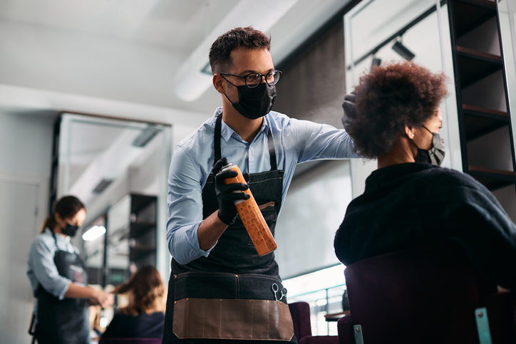 Cabeleireiro usando máscara facial protetora enquanto usa spray de cabelo e trabalha no cabelo de mulher negra.