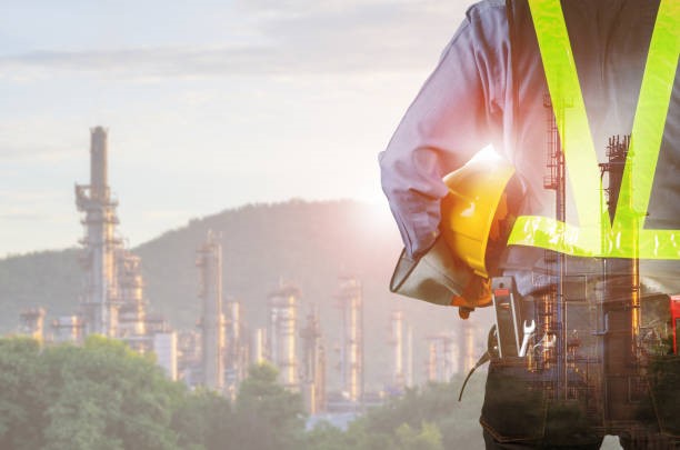 Dupla exposição do engenheiro por trás com ferramenta de sobrecarga segurando capacete amarelo para segurança dos trabalhadores, fundo de refinaria de petróleo turva.