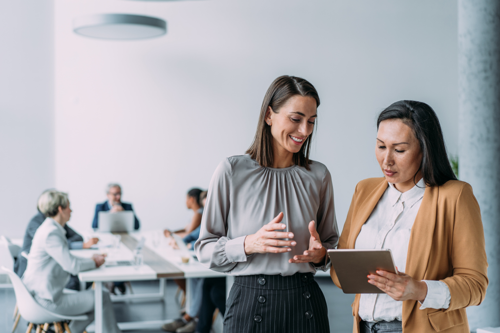 Duas mulheres conversam em um ambiente corporativo. Uma delas segura um tablet enquanto a outra sorri e gesticula